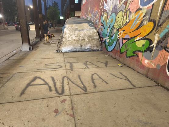 Outside of a tent under a bridge, someone has spray painted on the sidewalk the words: stay away.